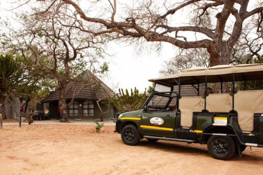 4 Day Kruger Park / Manyeleti Tour Wits Bushcamp Lodge A safari vehicle with canvas seats parked on a dirt path near a lodge. Bare trees and lush plants surround the area, evoking a serene, rustic atmosphere.