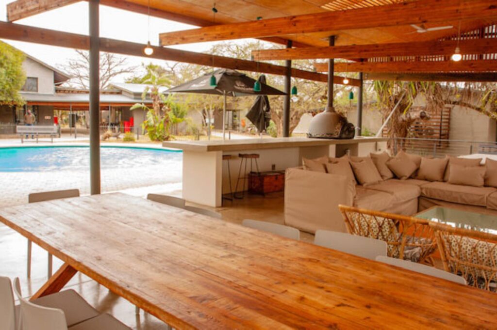 3 Day Kruger Park Covered patio with wooden tables and cushioned seating overlooks a pool. Umbrella and greenery create a relaxed, inviting outdoor atmosphere.