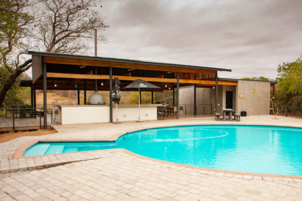 5 Day Kruger Park / Manyeleti Tour Wits Bushcamp Lodge Outdoor pool scene with a curved turquoise swimming pool, surrounded by gray pavers. A shaded bar area with tables is visible under a cloudy sky.