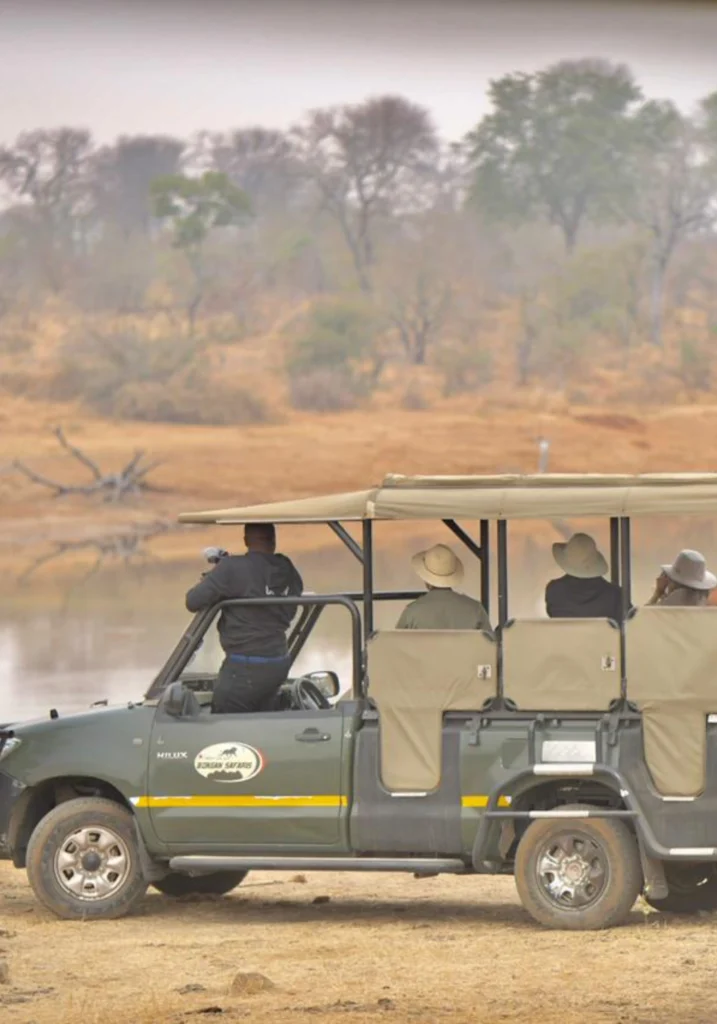 4 Day Kruger Park /Manyeleti tour African village Bongan lodge accommodation Open safari vehicle with tourists wearing hats, wildlife guide using binoculars, parked near a waterhole. Dry trees and hazy sky in the background.